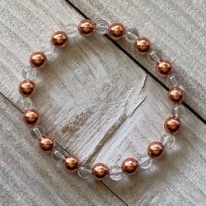 Pode incluir: Um bracelete feito de contas de cobre e cristal transparente alternadas. O bracelete est&aacute; sobre uma superf&iacute;cie de madeira branca.