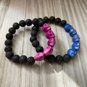 Könnte beinhalten: Zwei Armbänder aus schwarzen Lavasteinen mit Schädelperlen. Ein Armband hat rosa Schädelperlen und das andere blaue Schädelperlen.