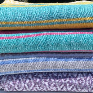 May include: A stack of four folded woven blankets. The top blanket is turquoise with yellow stripes. The second is turquoise with pink stripes. The third is white with light blue and grey stripes. The bottom blanket is purple with a white geometric pattern.