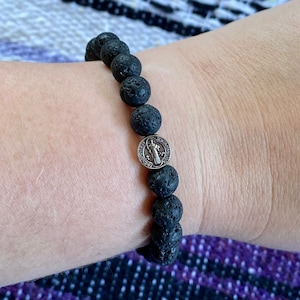 Puede incluir: Un brazalete de piedra de lava negra con un dije de San Miguel en plata. El brazalete está en la muñeca de una persona.