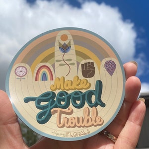 May include: A round sticker with a light blue background and a beige center. The sticker features a rainbow, a flower, a fist, and a heart. The text "Make Good Trouble" is written in a teal blue font. The sticker is made by feralFLORALS.