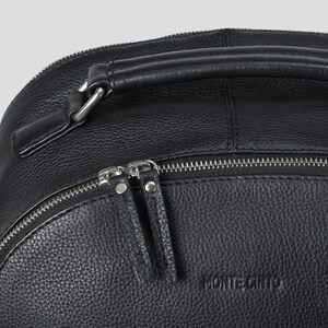 Puede incluir: Primer plano de una mochila de cuero negro con asa superior y cremalleras plateadas. La mochila tiene una superficie texturizada y la marca "MONTECRISTO" est&aacute; grabada en la parte delantera.