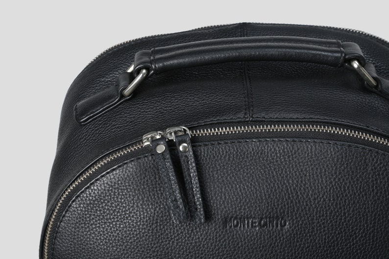 Puede incluir: Primer plano de una mochila de cuero negro con asa superior y cremalleras plateadas. La mochila tiene una superficie texturizada y la marca "MONTECRISTO" est&aacute; grabada en la parte delantera.