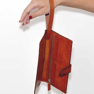 May include: A russet leather wallet with a wrist strap. The wallet is open, showing interior compartments and a zipped pocket. The strap is attached to the top of the wallet. The wallet has a buckle closure.
