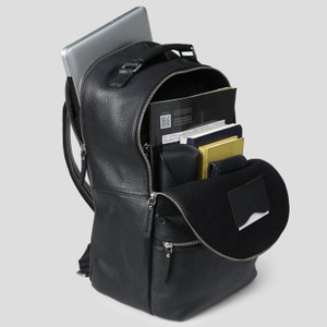 Puede incluir: Mochila de cuero negro con asa superior y correas ajustables. La mochila est&aacute; abierta, revelando un port&aacute;til, cuadernos y otros art&iacute;culos. La bolsa tiene m&uacute;ltiples compartimentos con cremallera y un acabado texturizado.