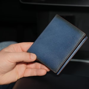 Puede incluir: Una cartera de cuero azul con un aspecto desgastado y envejecido. La cartera está doblada por la mitad y tiene un borde cosido.