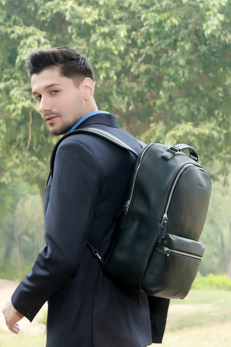 Puede incluir: Una mochila de cuero negro con cremalleras plateadas y un bolsillo frontal. La mochila se lleva sobre el hombro de una persona que lleva un traje de raya diplom&aacute;tica azul marino y una camisa azul. El fondo es una escena exterior verde borrosa.