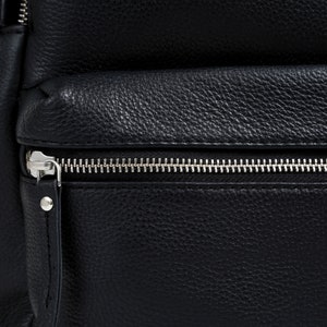 Puede incluir: Primer plano de una mochila de cuero negro con una superficie texturizada. La mochila tiene un cierre de cremallera plateada en el compartimento principal y un bolsillo frontal m&aacute;s peque&ntilde;o. El cuero tiene un grano granulado.