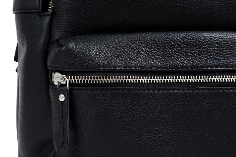 Puede incluir: Primer plano de una mochila de cuero negro con una superficie texturizada. La mochila tiene un cierre de cremallera plateada en el compartimento principal y un bolsillo frontal m&aacute;s peque&ntilde;o. El cuero tiene un grano granulado.