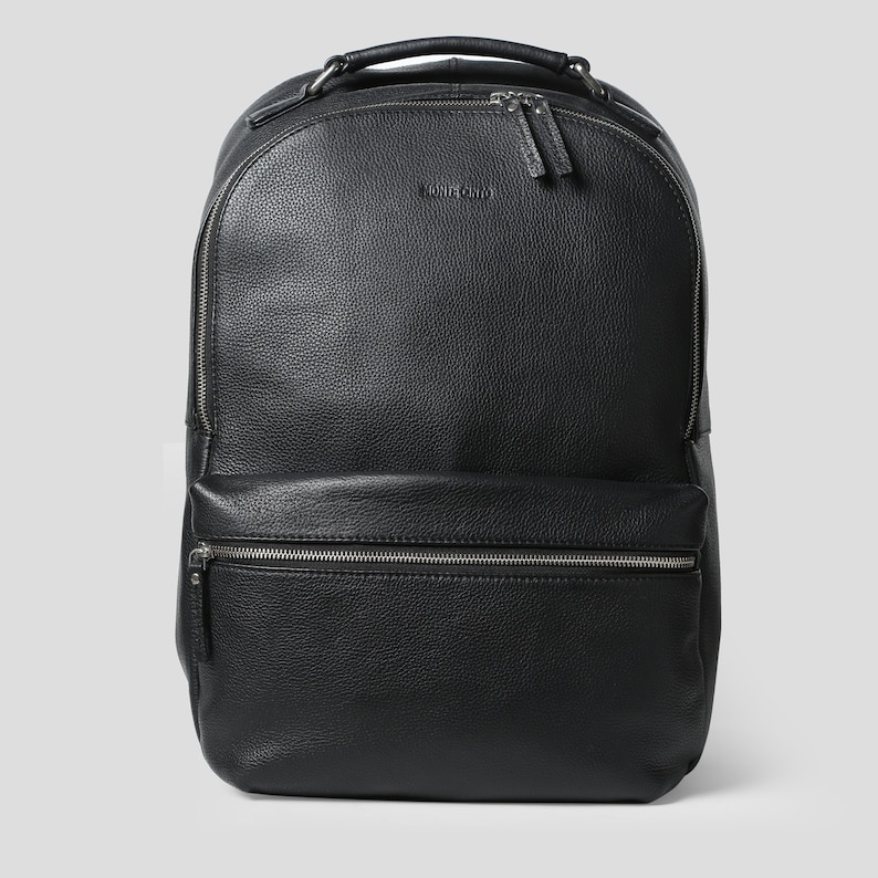 Puede incluir: Mochila de cuero negro con asa superior y m&uacute;ltiples compartimentos con cremallera. La mochila tiene un bolsillo frontal y cremalleras plateadas. La marca "MONTE ORIO" es visible en la parte delantera.