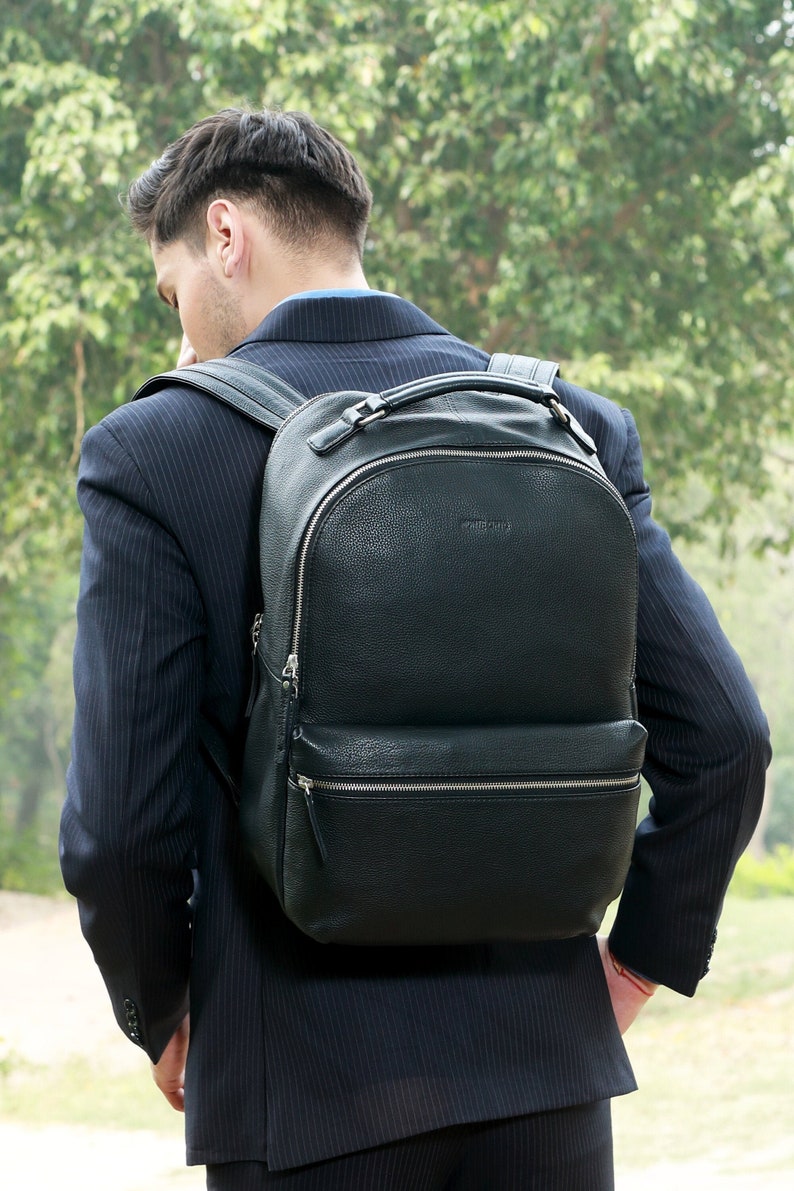 Puede incluir: Una mochila de cuero negro con la parte superior redondeada y un bolsillo frontal con cremallera. La mochila tiene un asa superior y correas ajustables para los hombros. La imagen muestra la mochila sobre una persona que lleva un traje oscuro.