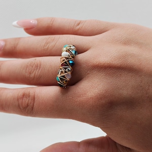 May include: A handmade ring with a gold wire wrap design. The ring features a variety of colorful beads, including turquoise, red, white, and blue. The beads are arranged in a vertical pattern, creating a unique and eye-catching accessory.