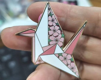 Può includere: Uno spillo a forma di gru origami dorata con dettagli floreali rosa e verdi. La gru è piegata in uno stile tradizionale e ha uno sfondo bianco.