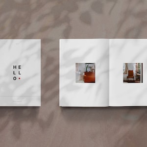 Könnte beinhalten: Ein weißes Buch mit dem Wort "HELLO" in schwarzer Schrift und einem roten Punkt. Das Buch ist geöffnet und zeigt zwei Innenseiten mit einem kleinen quadratischen Fensterausschnitt auf jeder Seite. Die Fenster zeigen einen Blick in einen Raum mit Möbeln.