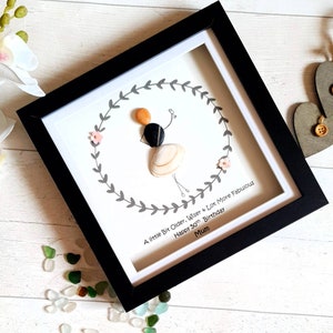 Pu&ograve; includere: Un'opera d'arte incorniciata con una figura di sassi che tiene dei palloncini, circondata da una ghirlanda grigia e fiori rosa. La cornice nera circonda un passe-partout bianco con il testo: "A little Bit Older, Wiser + Lot More Fabulous Happy 50th Birthday Mum."