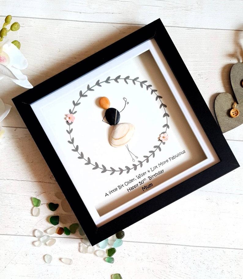 Pu&ograve; includere: Un'opera d'arte incorniciata con una figura di sassi che tiene dei palloncini, circondata da una ghirlanda grigia e fiori rosa. La cornice nera circonda un passe-partout bianco con il testo: "A little Bit Older, Wiser + Lot More Fabulous Happy 50th Birthday Mum."