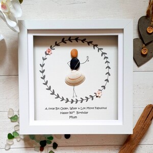 Pu&ograve; includere: Un'opera d'arte incorniciata con una figura di sassi all'interno di una ghirlanda. La figura &egrave; fatta di sassi e una conchiglia, che tiene in mano un drink. Il testo recita: "A little Bit Older, Wiser & Lot More Fabulous Happy 50th Birthday Mum."