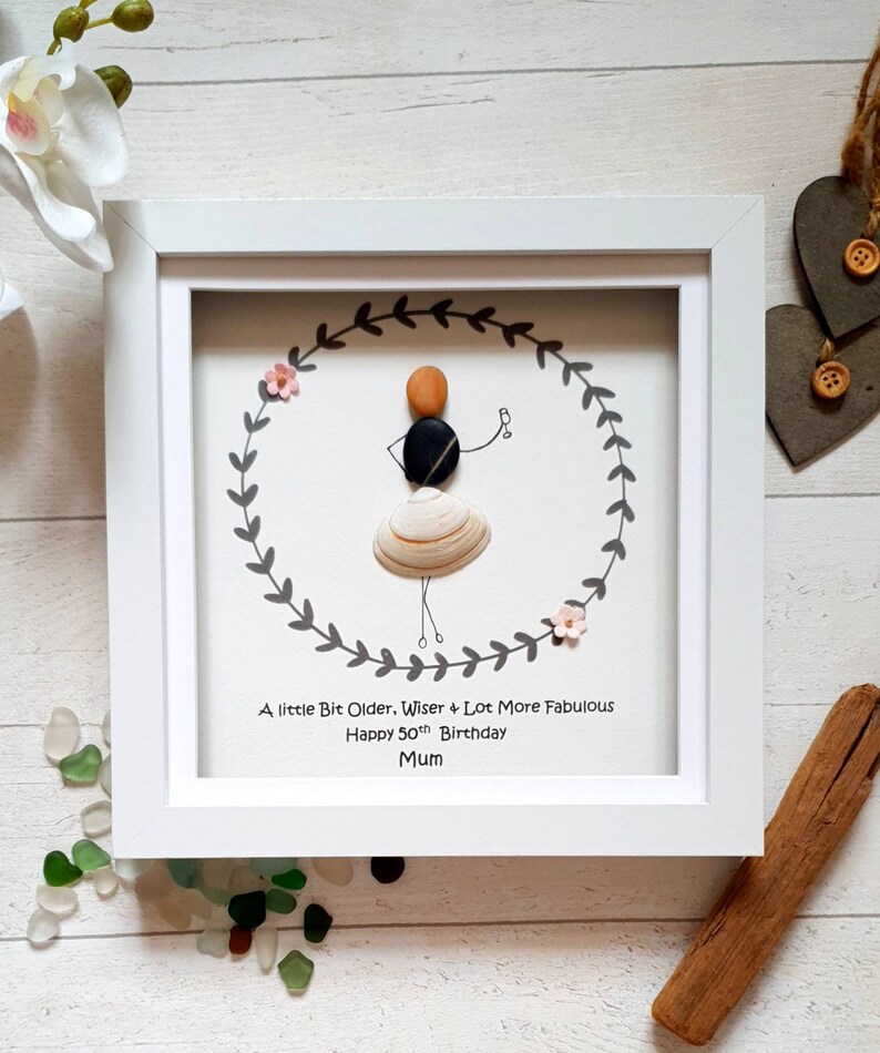 Pu&ograve; includere: Un'opera d'arte incorniciata con una figura di sassi all'interno di una ghirlanda. La figura &egrave; fatta di sassi e una conchiglia, che tiene in mano un drink. Il testo recita: "A little Bit Older, Wiser & Lot More Fabulous Happy 50th Birthday Mum."