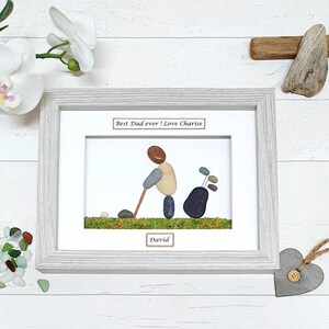 May include: Framed pebble art depicting a golfer with a golf bag. The artwork features a light gray wooden frame, white matting, and the text "Best Dad ever! Love Charise" and "David". The golf scene is created with pebbles and green moss.