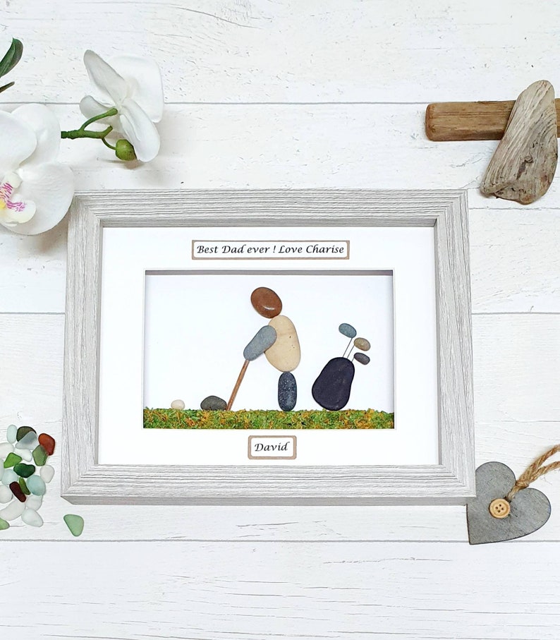 May include: Framed pebble art depicting a golfer with a golf bag. The artwork features a light gray wooden frame, white matting, and the text "Best Dad ever! Love Charise" and "David". The golf scene is created with pebbles and green moss.