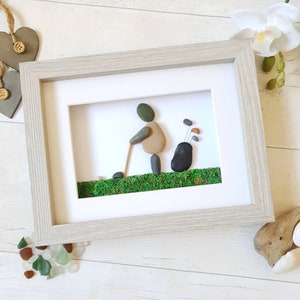 May include: Framed pebble art depicting a golfer in action, crafted with smooth stones. The artwork features a golfer with a golf club, a golf bag, and green moss representing the course. The frame is a light gray color.