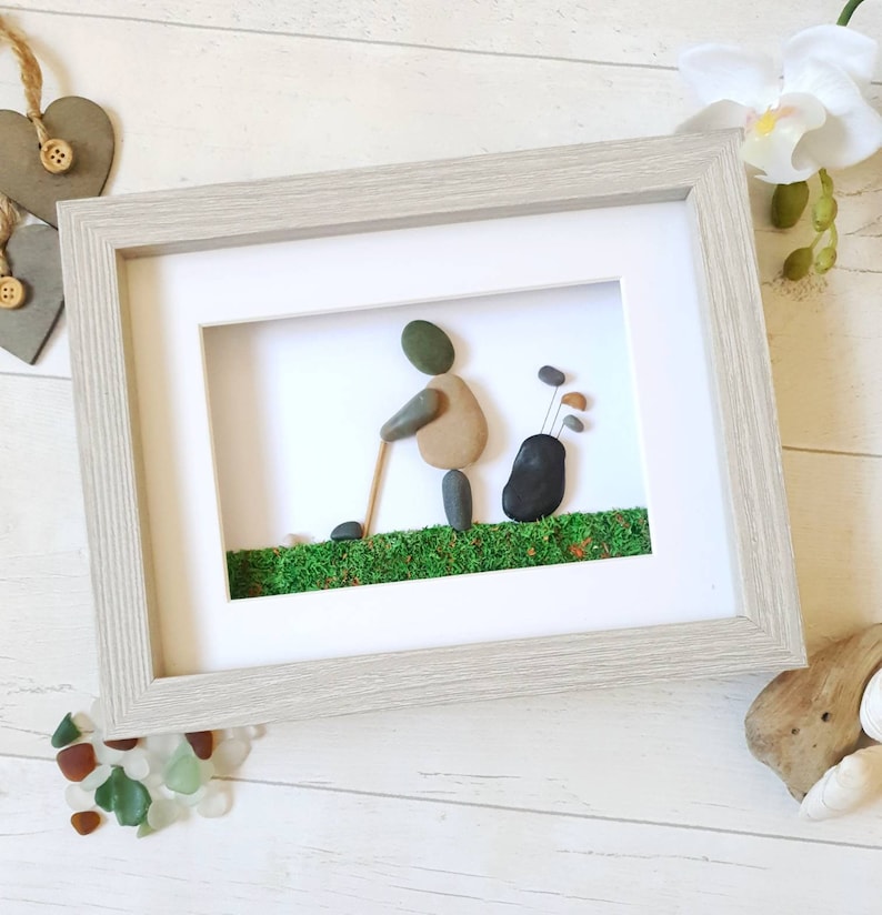 May include: Framed pebble art depicting a golfer in action, crafted with smooth stones. The artwork features a golfer with a golf club, a golf bag, and green moss representing the course. The frame is a light gray color.