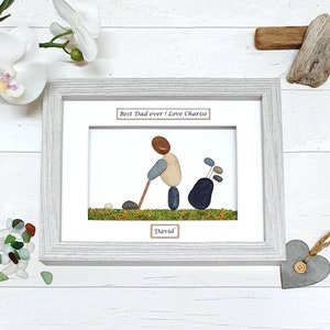 May include: Framed pebble art depicting a golfer with a golf bag. The artwork features a golfer made of pebbles, swinging a golf club. The text "Best Dad ever! Love Charise" and "David" are included. The frame is light gray.