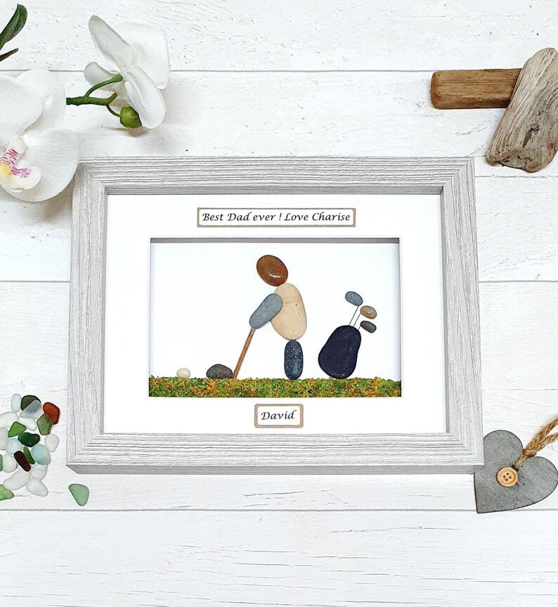 May include: Framed pebble art depicting a golfer with a golf bag. The artwork features a golfer made of pebbles, swinging a golf club. The text "Best Dad ever! Love Charise" and "David" are included. The frame is light gray.