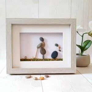 May include: Framed pebble art depicting a golfer with a golf bag. The artwork features a light gray wooden frame with a white mat. The golfer and golf bag are created from smooth, gray and brown pebbles. The background is white with a green base.
