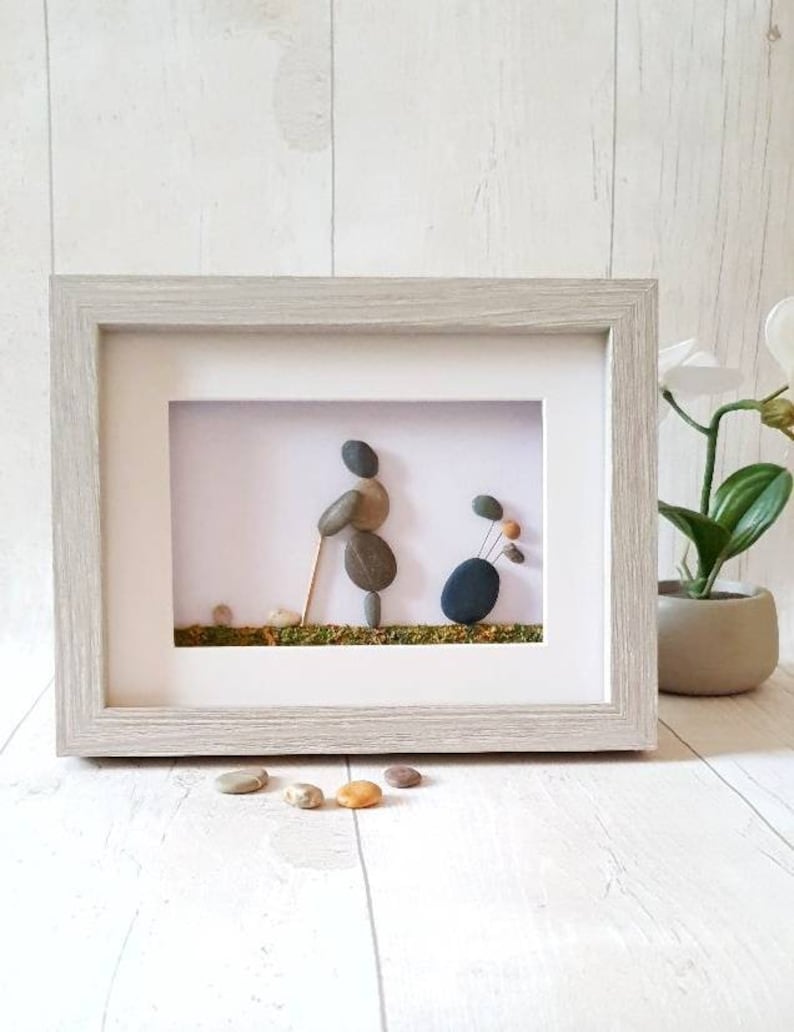 May include: Framed pebble art depicting a golfer with a golf bag. The artwork features a light gray wooden frame with a white mat. The golfer and golf bag are created from smooth, gray and brown pebbles. The background is white with a green base.