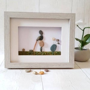 May include: Framed pebble art depicting a golfer in action, swinging a club. The artwork features a light gray wooden frame, with the golfer and golf clubs crafted from smooth, natural stones. The background includes a green grassy area.