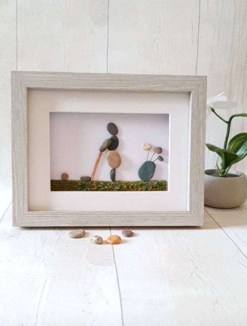 May include: Framed pebble art depicting a golfer in action, swinging a club. The artwork features a light gray wooden frame, with the golfer and golf clubs crafted from smooth, natural stones. The background includes a green grassy area.
