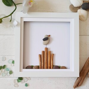 以下が含まれることがあります： 鳥が棒の積み重ねの上に止まっている、石で作った額装されたアート作品。鳥は灰色と黒の石でできており、棒は茶色です。背景は白です。