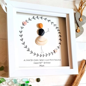 Pu&ograve; includere: Un'opera d'arte con sassi incorniciata in una cornice bianca. L'arte presenta una figura di sassi all'interno di una ghirlanda di foglie grigie, con il testo: "A little Bit Older, Wiser & Lot More Fabulous Happy 50th Birthday Mum."