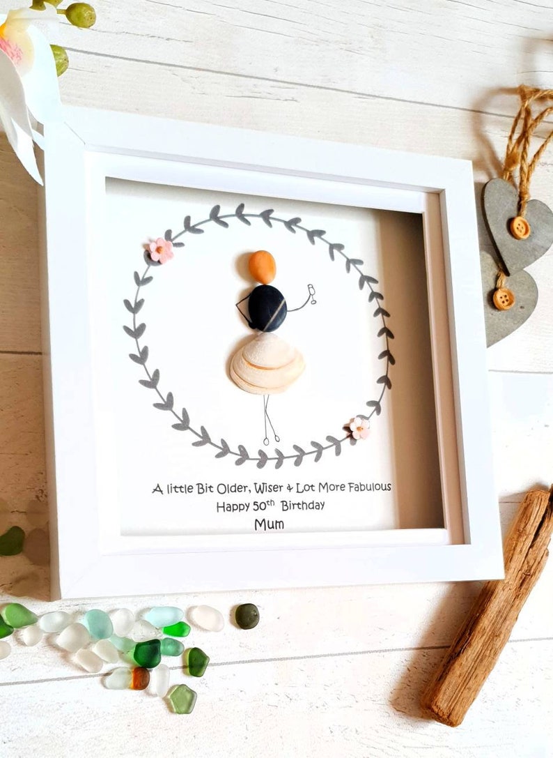 Pu&ograve; includere: Un'opera d'arte con sassi incorniciata in una cornice bianca. L'arte presenta una figura di sassi all'interno di una ghirlanda di foglie grigie, con il testo: "A little Bit Older, Wiser & Lot More Fabulous Happy 50th Birthday Mum."