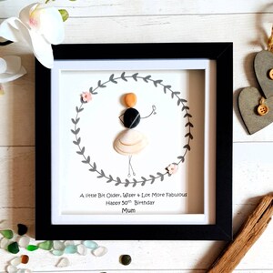 Pu&ograve; includere: Opera d'arte incorniciata con una figura di ciottoli all'interno di una ghirlanda di foglie grigie. La figura &egrave; fatta di ciottoli e conchiglie, con il testo "A little Bit Older, Wiser & Lot More Fabulous Happy 50th Birthday Mum". La cornice &egrave; nera.