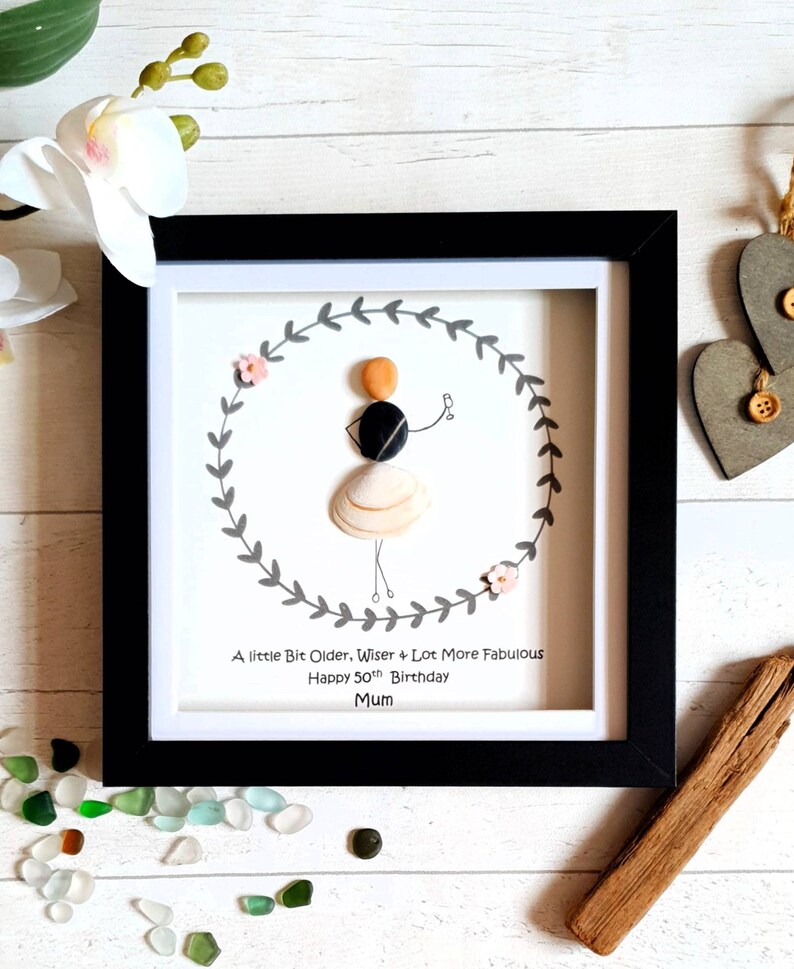 Pu&ograve; includere: Opera d'arte incorniciata con una figura di ciottoli all'interno di una ghirlanda di foglie grigie. La figura &egrave; fatta di ciottoli e conchiglie, con il testo "A little Bit Older, Wiser & Lot More Fabulous Happy 50th Birthday Mum". La cornice &egrave; nera.