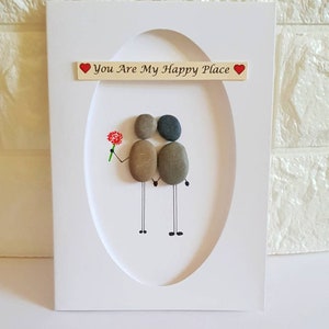 May include: White card with a framed pebble art design of two figures holding hands. The text "You Are My Happy Place" is written on the card with a red heart on either side of the text.