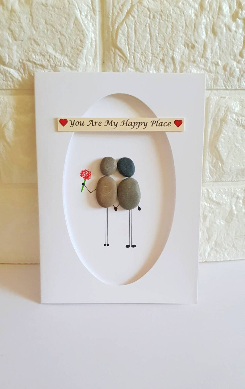 May include: White card with a framed pebble art design of two figures holding hands. The text "You Are My Happy Place" is written on the card with a red heart on either side of the text.