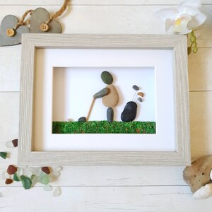May include: Framed pebble art depicting a golfer in action, crafted with natural stones. The artwork features a golfer with a golf club, a golf bag, and green moss representing the course, all within a light gray wooden frame.