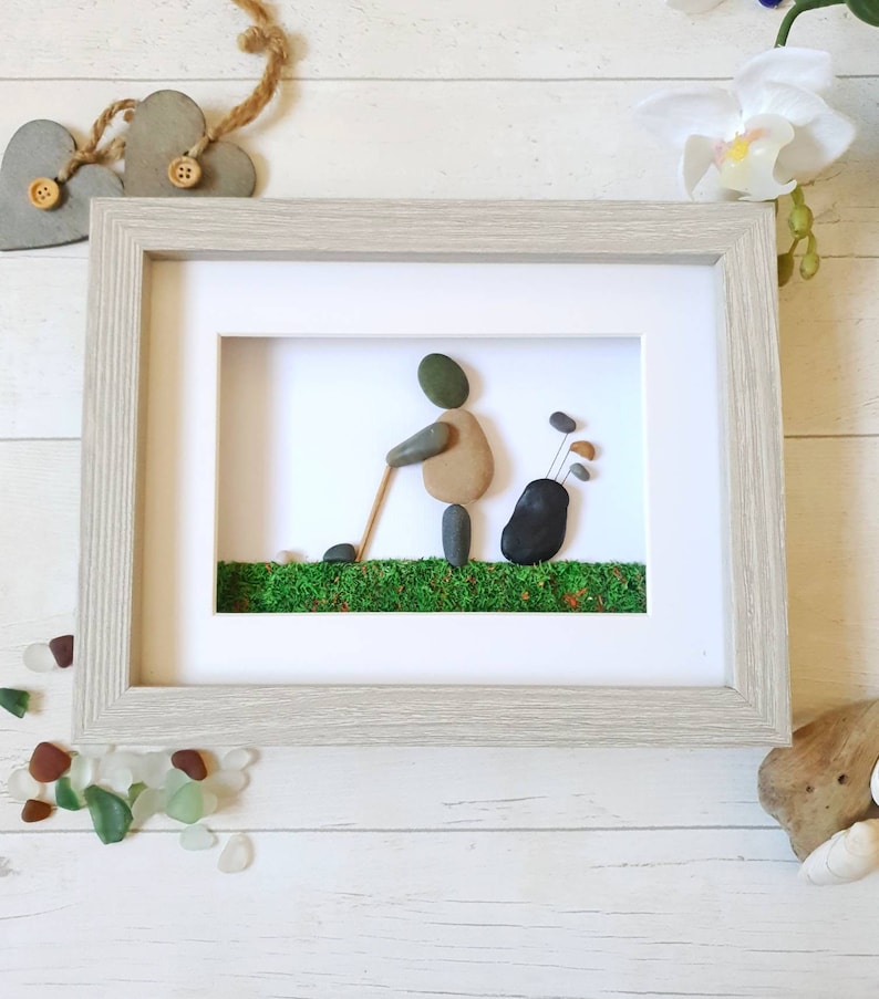 May include: Framed pebble art depicting a golfer in action, crafted with natural stones. The artwork features a golfer with a golf club, a golf bag, and green moss representing the course, all within a light gray wooden frame.