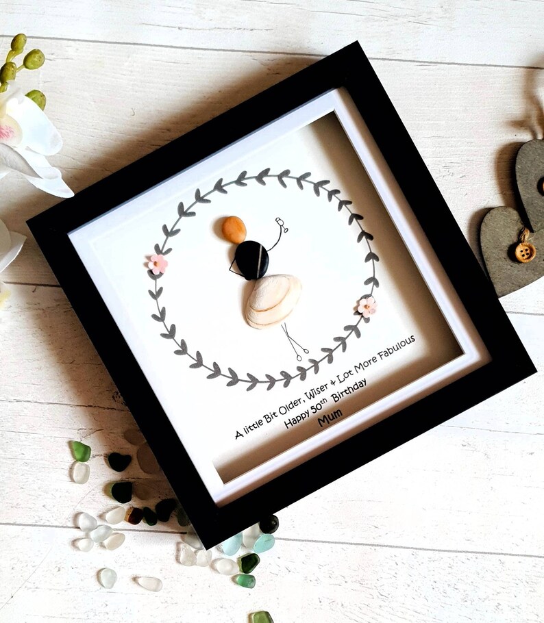 Pu&ograve; includere: Opera d'arte incorniciata con una figura fatta di conchiglie e pietre, che tiene un palloncino. La figura &egrave; circondata da una ghirlanda, con il testo: "A little Bit Older, Wiser + Lot More Fabulous Happy 50th Birthday Mum."