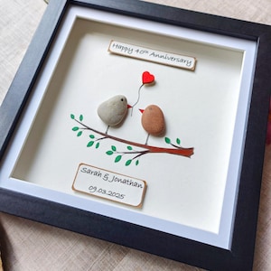 May include: A framed artwork featuring two painted pebble birds on a branch with green leaves. The birds are holding a red heart. The frame has a white mount and a black wood frame. The artwork includes the text "Happy 40th Anniversary", "Sarah & Jonathan", and "09.03.2025".
