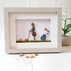 May include: Framed pebble art depicting a golfer in action, with a golf bag and ball. The artwork features a light gray wooden frame. The golfer is made of brown and gray pebbles, set against a white background with green moss.