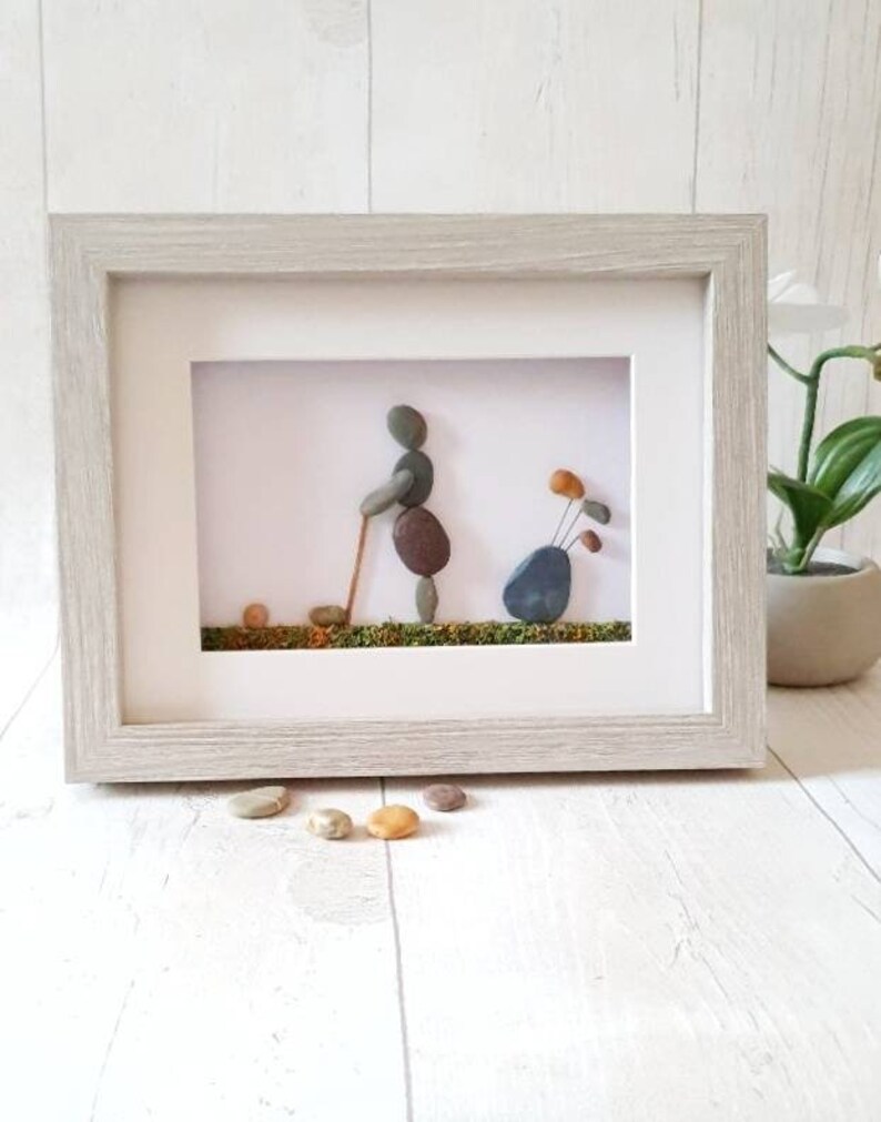 May include: Framed pebble art depicting a golfer in action, with a golf bag and ball. The artwork features a light gray wooden frame. The golfer is made of brown and gray pebbles, set against a white background with green moss.
