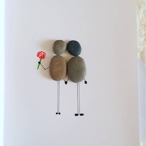May include: A handmade greeting card with a couple made from small stones. The couple is holding hands and the one on the left is holding a red flower. The card is white with black lines drawn on it.
