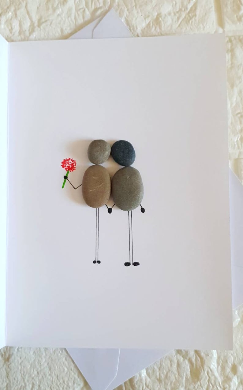 May include: A handmade greeting card with a couple made from small stones. The couple is holding hands and the one on the left is holding a red flower. The card is white with black lines drawn on it.