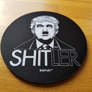 May include: A black round mousepad with a white illustration of a person with a mustache and the word "SHITLER" in white letters. The text "DabPadz™" is also in white letters at the bottom of the mousepad.