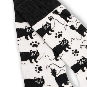 May include: A pair of white socks with a black cat and paw print pattern. The socks have black heels, toes, and cuffs. The cat design includes a playful string.