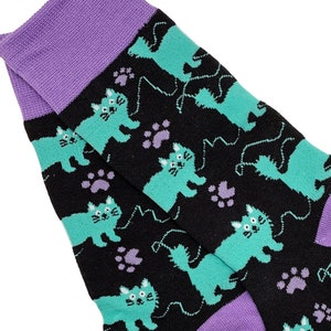 Op de afbeelding: Een paar zwarte sokken met een paarse boord. De sokken hebben een herhalend patroon van turquoise katten met zwart garen en roze pootjes.